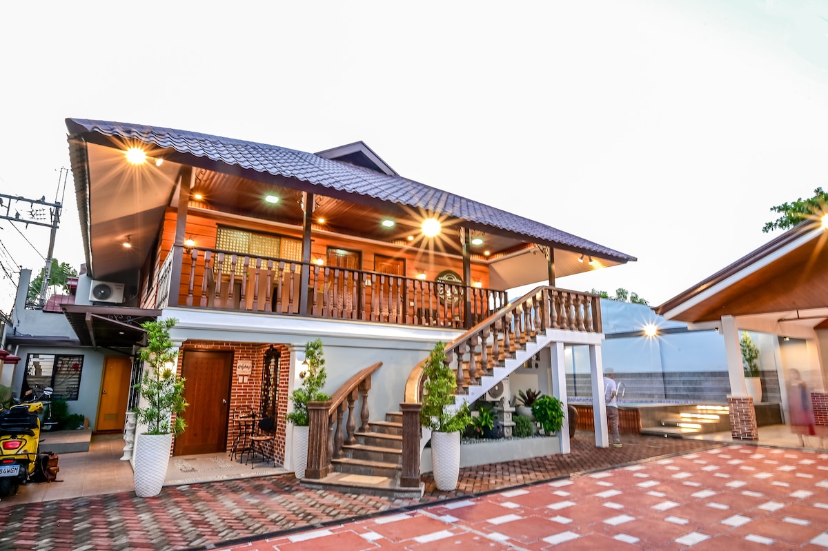 A traditional two-story Filipino-style cabin features a charming wooden exterior and a welcoming balcony. Well-lit with outdoor lighting, the entrance includes a staircase leading to the upper level, surrounded by neatly arranged plants in pots. The surrounding area is paved with patterned bricks.
