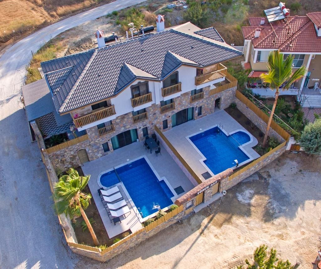 An aerial view showcases a spacious villa with an inviting stone façade and two private swimming pools. Surrounding gardens and lounge chairs are visible beside each pool, emphasizing the outdoor relaxation areas. The villa is situated in a tranquil environment with nearby structures.