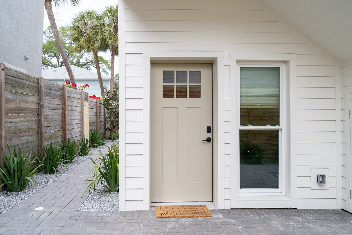 The entrance to the villa features a light beige door with four small windows at the top. A welcome mat is placed at the base of the door. Adjacent to the door is a large window, allowing natural light to fill the entryway. Lush greenery lines the pathway leading to the entrance.