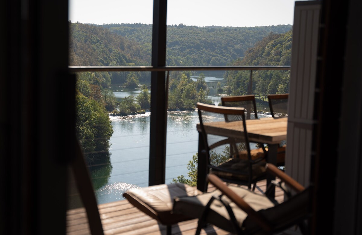 The image captures a serene view of the river and surrounding greenery through large glass doors. A wooden terrace is visible, featuring a table and chairs overlooking the tranquil waters, inviting leisurely relaxation amidst nature.
