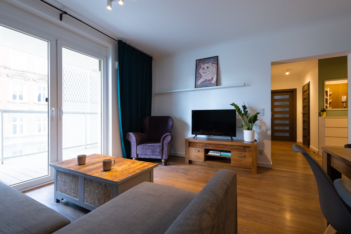 A spacious living area is showcased, featuring a comfortable sofa and a warm wooden coffee table. A television is positioned on a side cabinet, while natural light filters in through sliding glass doors that open to a balcony. Decorative elements include a potted plant and art on the wall.