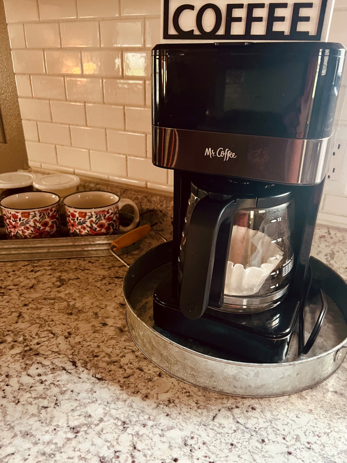 A coffee maker sits on a polished granite countertop, featuring a glass carafe and a filter basket. Three decorative mugs are arranged on a tray nearby, displaying a floral design against a backdrop of white tiled backsplash.
