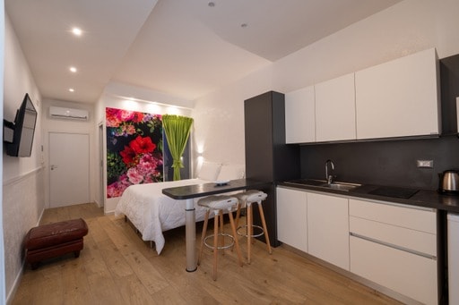 A bright studio apartment features a bed against a vibrant floral wall, enhancing the space's modern design. A compact kitchen is equipped with essential appliances, while a small dining area with two stools offers seating. Warm wood flooring adds a cozy touch to the overall aesthetic.