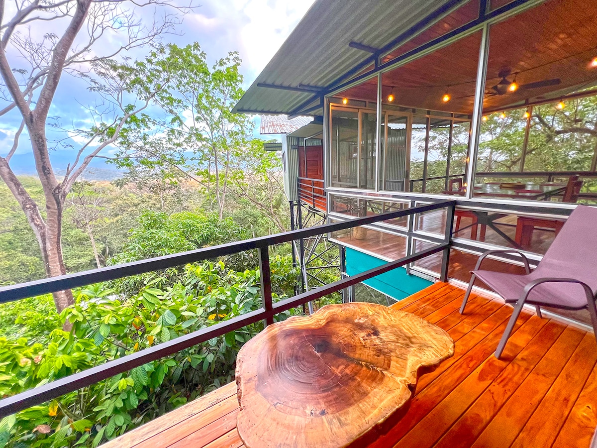 A wooden deck showcases a large slab table, surrounded by greenery and expansive mountain views. Glass walls create a connection between the outdoor space and the interior, which is illuminated by string lights overhead.
