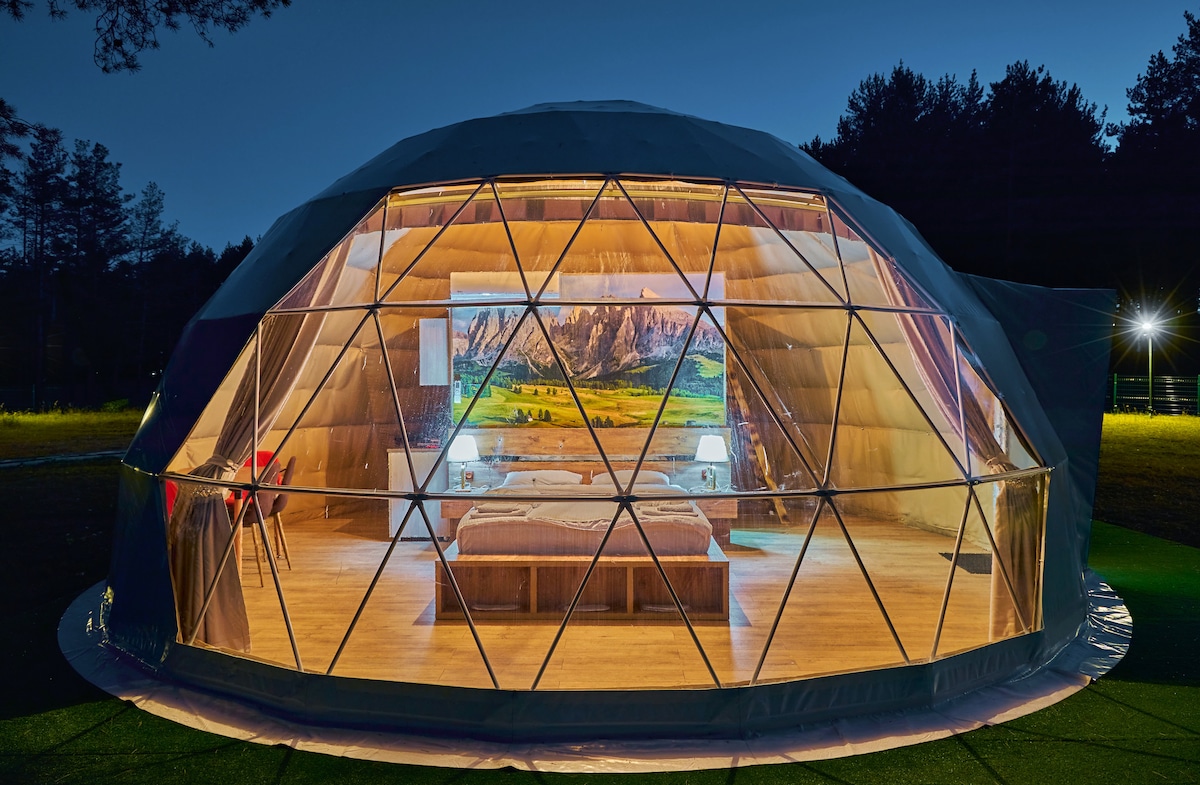 A geodesic dome structure is illuminated at night, showcasing a spacious interior with large windows. The warm lighting highlights two beds and a scenic mural of mountains. A dining area with chairs is visible, enhancing the inviting space surrounded by nature.