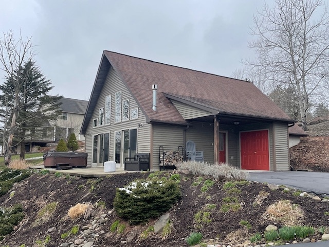 The exterior of a modern-style home features large windows that allow natural light to fill the interior. A welcoming red door stands out against neutral siding. A spacious deck with a hot tub overlooks landscaped areas with greenery and seasonal foliage, enhancing the outdoor space.