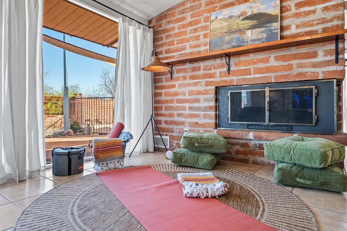 A tranquil corner of the living room features a circular jute rug with a yoga mat placed on it. Green floor cushions are arranged nearby, alongside a cozy blanket. Natural light streams in from large sliding doors, which open to reveal a scenic outdoor view.