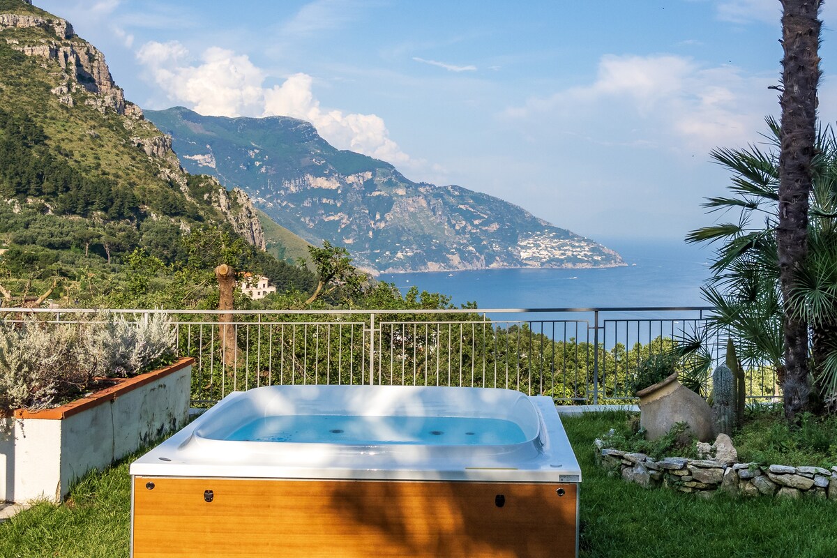 A relaxing outdoor area features a hot tub surrounded by lush greenery, with a panoramic view of the coastline and hills in the background. The scene captures a serene atmosphere, highlighting the natural beauty of the Amalfi Coast.