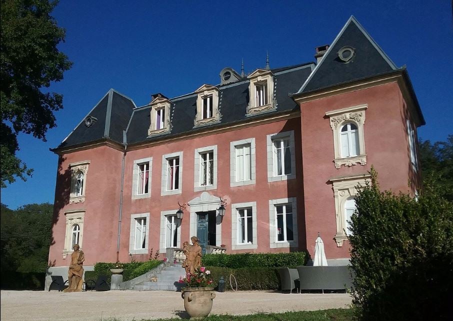 The large pink manor house features a symmetrical façade with decorative stone details and multiple windows. A set of stairs leads to the main entrance, flanked by two stone statues. Lush greenery and neatly trimmed bushes surround the property, with a clear blue sky above.