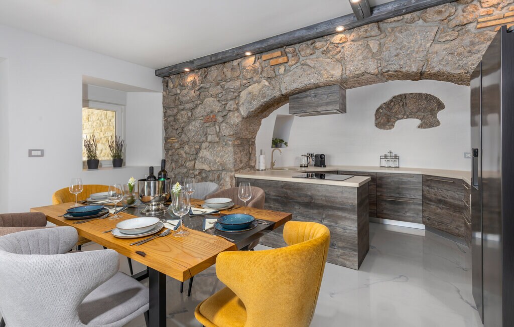 A modern kitchen area features a rustic stone arch and sleek cabinetry. A wooden dining table is set with plates, glasses, and cutlery, accompanied by colorful chairs. Soft lighting enhances the stone wall, while a large window offers natural light into the space.