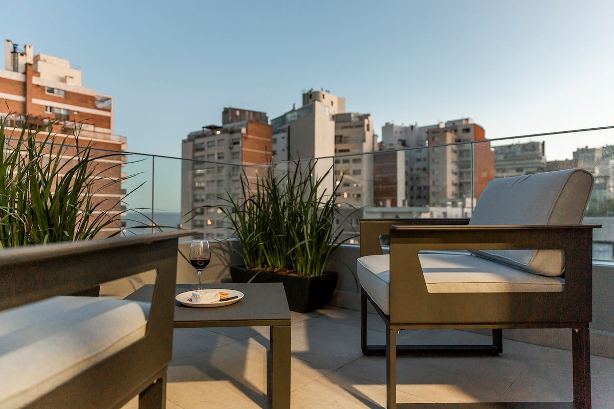 An outdoor balcony is shown, featuring a small table with a plate and a glass, surrounded by modern seating. Green plants are placed in the corners, and a city skyline is visible in the background, reflecting the evening light.