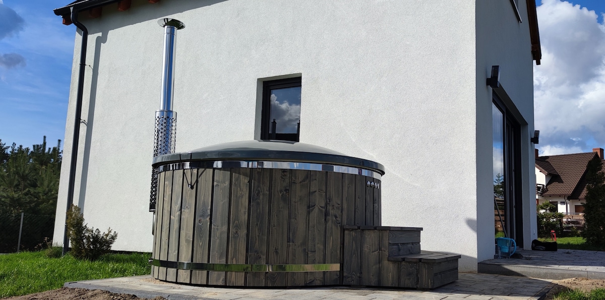 A wooden hot tub is situated outside the house, featuring a circular design and a sleek cover. Steps lead up to the tub, while a chimney is visible nearby, indicating a heat source. The exterior wall of the house is painted white, complementing the natural surroundings.