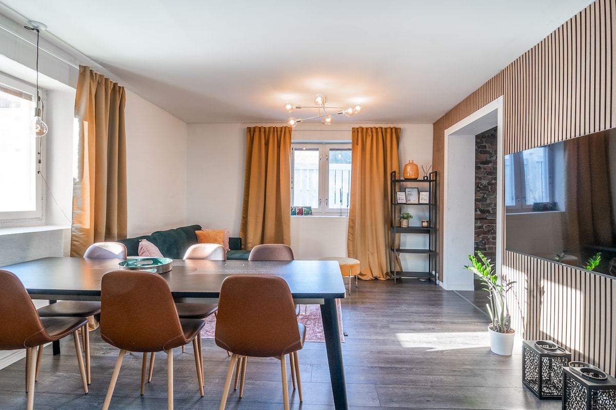 A spacious living area is featured, showcasing a dining table surrounded by several wooden chairs. Natural light enters through large windows, partially covered by flowy curtains. A green sofa and decorative shelf add character to the space, which is illuminated by modern light fixtures.