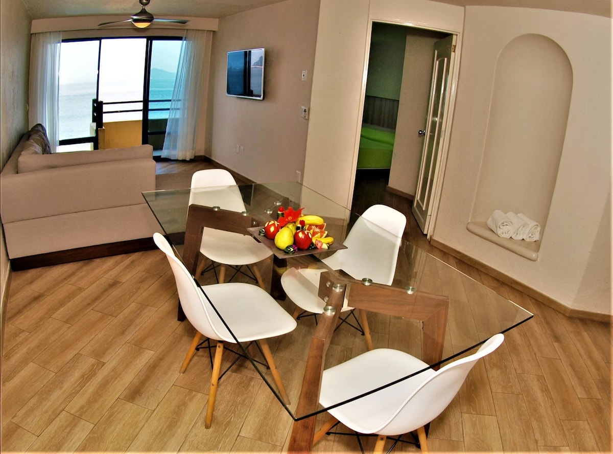 A bright dining area features a glass-top table surrounded by four white chairs. A bowl of fresh fruit is centrally placed. A comfortable sofa and a wall-mounted television are visible in the background, with large windows showcasing ocean views.
