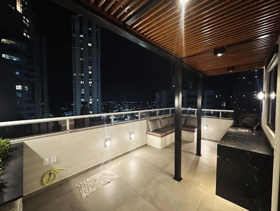 An open rooftop space is shown, featuring a seating area with soft cushions positioned against the wall. Ambient lighting is installed, highlighting the modern design in the evening. The city skyline is visible in the background, creating a serene atmosphere.
