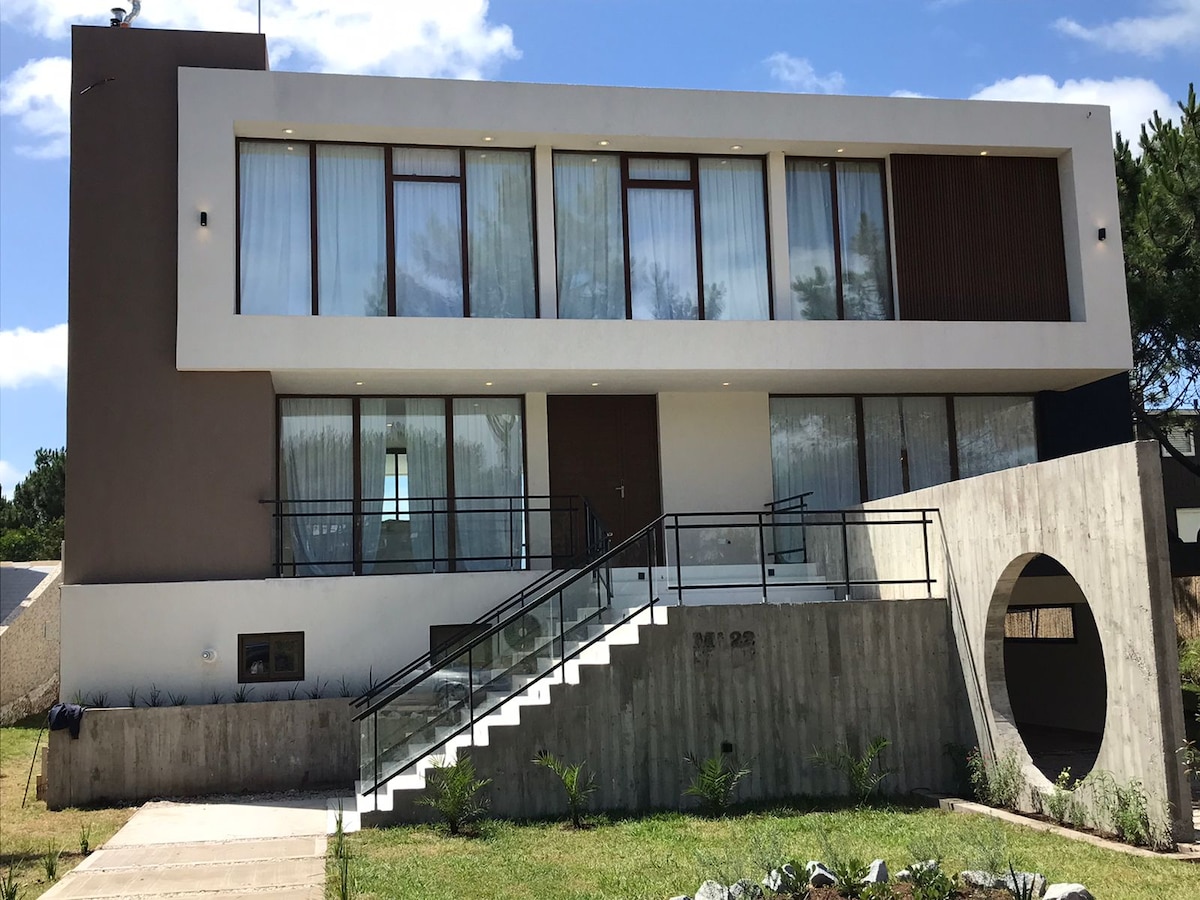 A contemporary two-story building is showcased, featuring large glass windows that allow natural light to fill the interior. A welcoming entrance is highlighted by a ramp, ensuring accessibility. Lush greenery and small ornamental plants adorn the exterior, contributing to a serene environment.