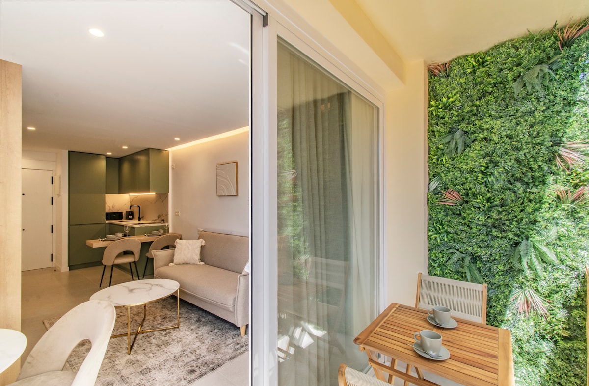 An inviting terrace features a small wooden table with two chairs positioned against a lush green wall of plants. Through large glass doors, a modern living area is visible, showcasing soft seating and a well-equipped kitchen area.