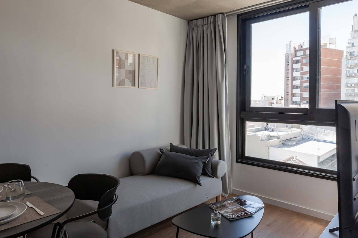 A living area features a modern sofa in light grey, positioned near a large window with views of the cityscape. A round coffee table lies in front of the sofa, accompanied by a dining table set for four, enhancing the space's functionality.