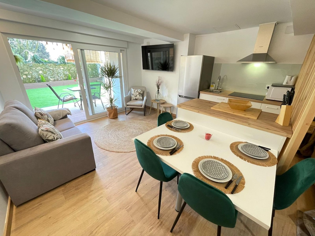 A modern open-plan living area features a cozy gray sofa and a dining table set with place settings for four. Large glass doors provide a view of a private garden area, while a small rug sits beneath a round dining table. A flat-screen television is mounted on the wall.