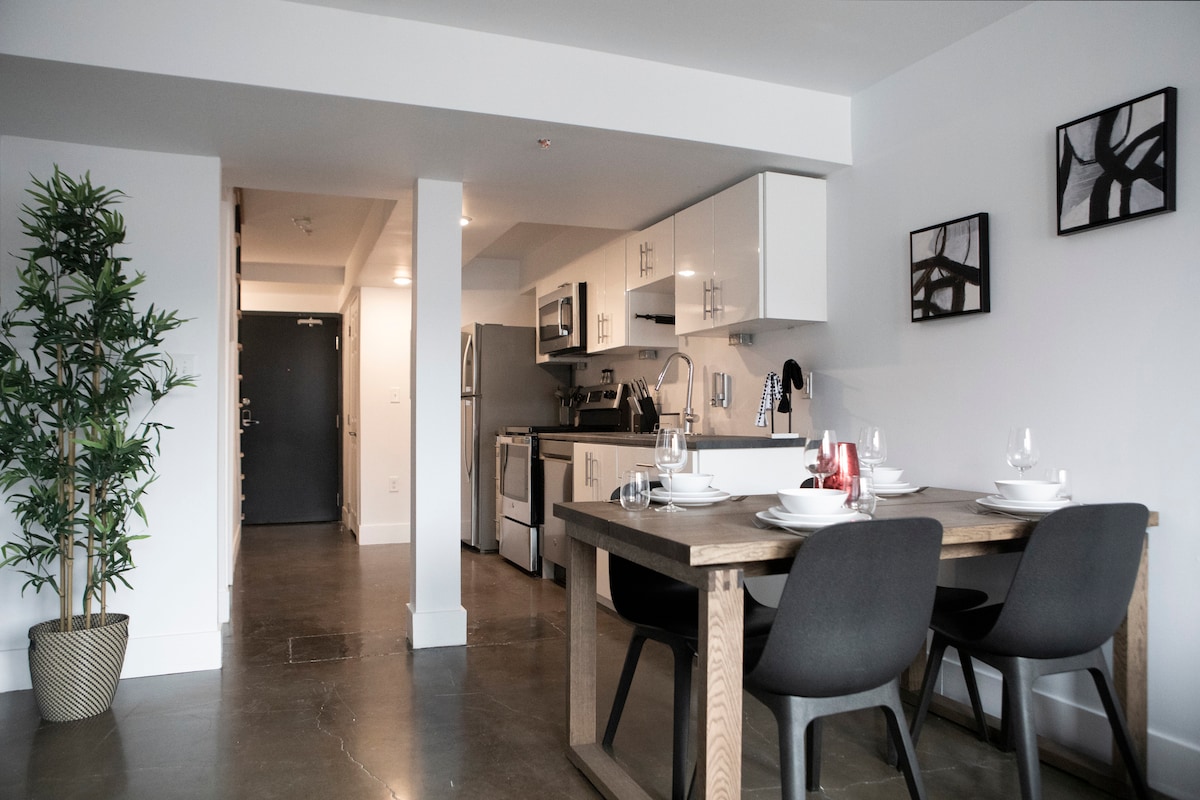 A modern dining area features a wooden table set for four, complemented by sleek black chairs. The kitchen is visible in the background, showcasing stainless steel appliances and minimalist cabinetry. A potted plant adds a touch of greenery, while wall art decorates the space.