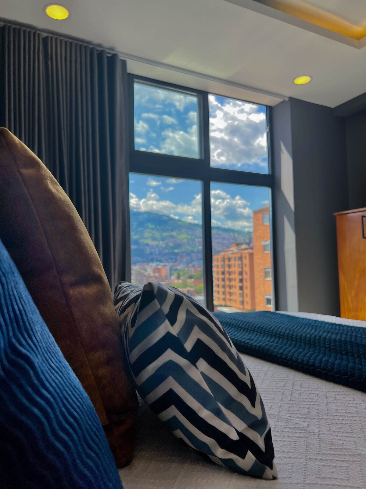 A cozy bedroom features a view of the mountains and sky through large modern windows. The space is adorned with decorative pillows in rich textures, positioned on a plush bed. Natural light fills the room, enhancing its inviting ambiance.