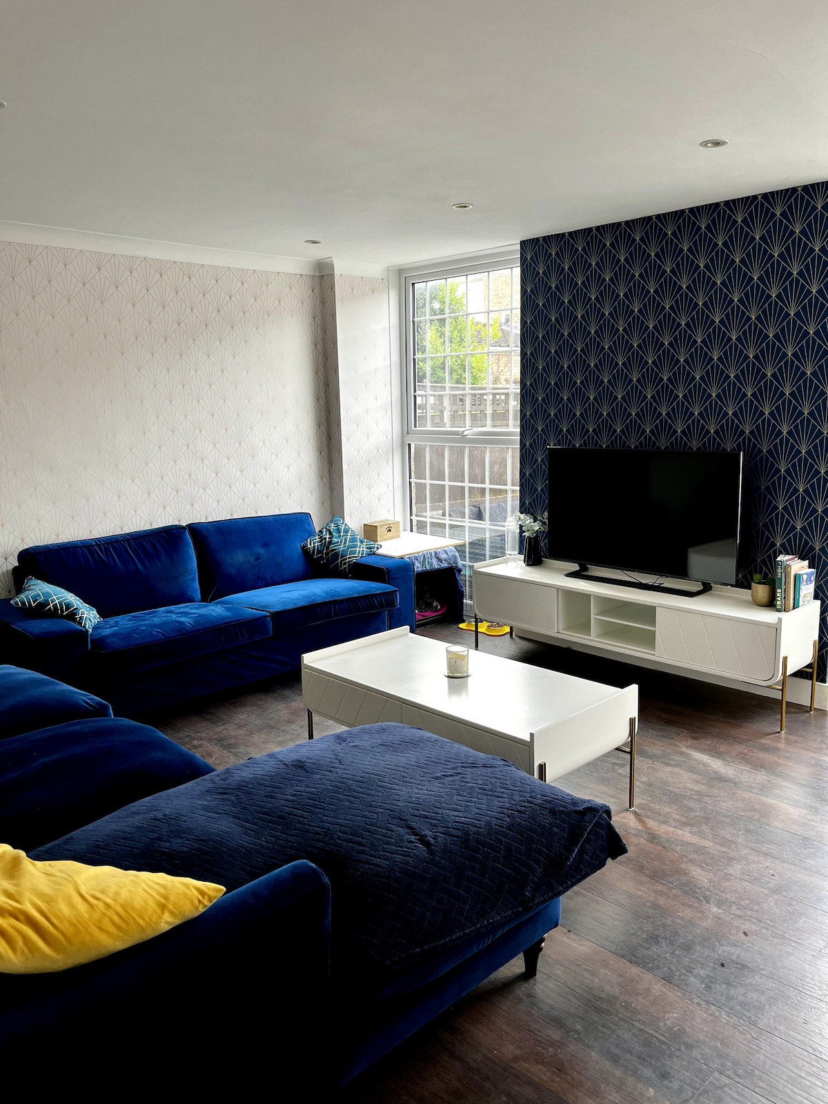 A spacious living room features a deep blue sectional sofa and coordinating accent pillows. A white coffee table is positioned in front of the sofa, with a flat-screen television mounted on a textured dark wall. Natural light streams in through a large window, complementing the decor.