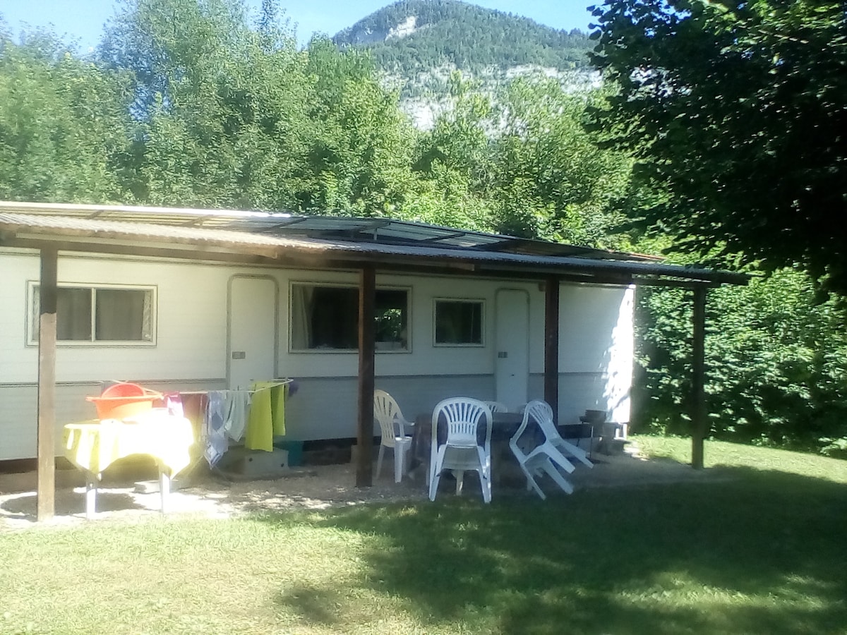 A mobile home is set within a grassy area surrounded by trees. Outdoor furniture, including several white chairs and a colorful table, is positioned in front. The roof features an overhang, providing shade. The mountains are visible in the background, adding to the natural ambiance.