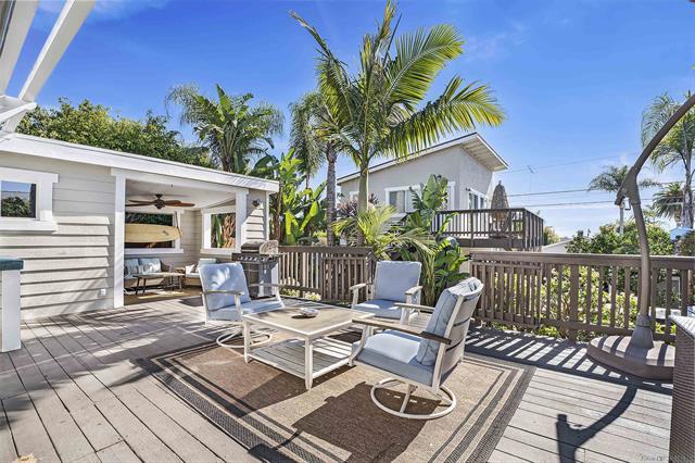 A spacious outdoor deck features comfortable seating with light-colored chairs and a circular table, surrounded by tropical plants. An overhead fan is visible, providing a cooling effect. Sunshine highlights the inviting atmosphere, enhancing the space for relaxation and social gatherings.