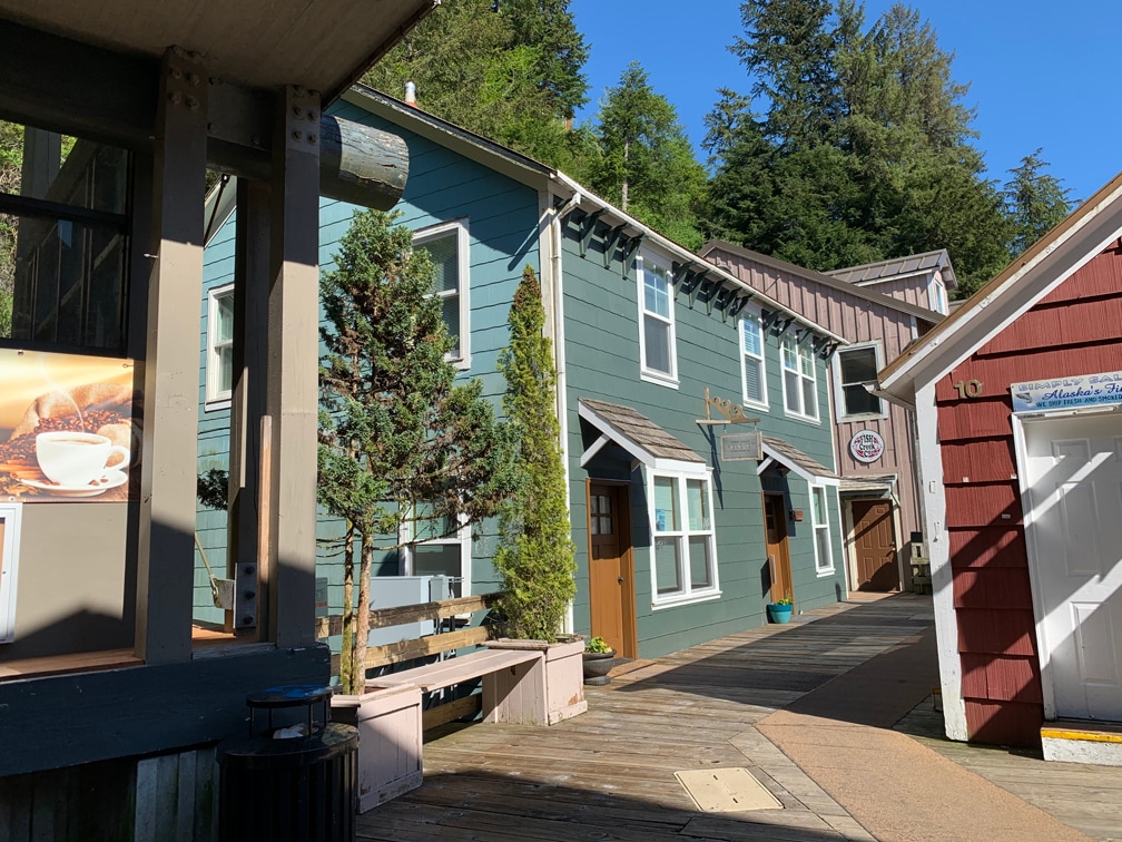A charming row of buildings is depicted, showcasing green and red exteriors. Wooden walkways connect the structures, with trees and shrubs providing natural greenery. The entrance to the suite is visible, complemented by large windows, inviting light into the environment.