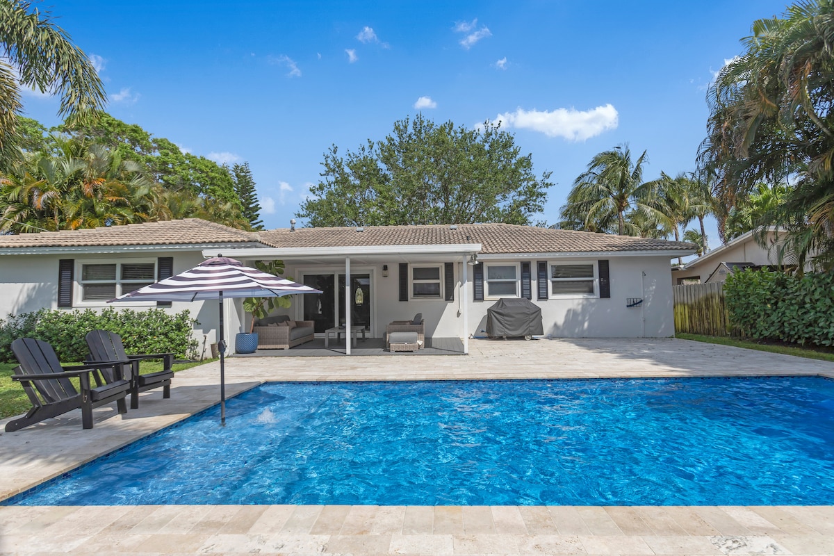 The outdoor area features a large pool surrounded by a stone deck, with comfortable seating beneath an umbrella. Lush greenery and palm trees create a serene backdrop, while the house provides ample shaded space for relaxation.