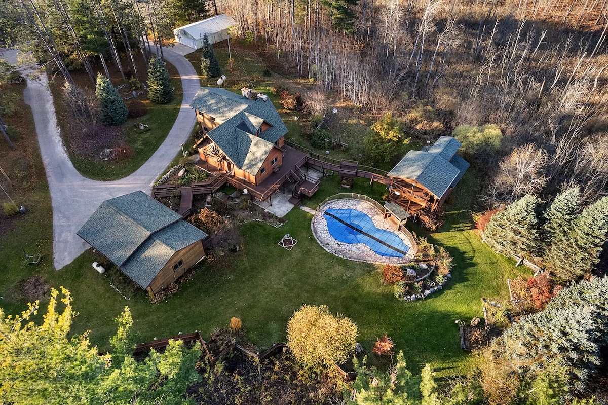 An aerial view captures the expansive property featuring two cabins surrounded by lush greenery. A spacious patio area and a heated inground pool are visible in the center, complemented by landscaped gardens. Tall trees and manicured lawns frame the serene setting.
