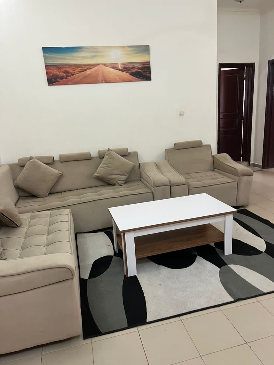 A modern living area is featured, showcasing a light-colored sectional sofa with multiple cushions. A simple white coffee table sits on a patterned rug, while a landscape artwork is displayed on the wall, contributing to a comfortable and inviting space.
