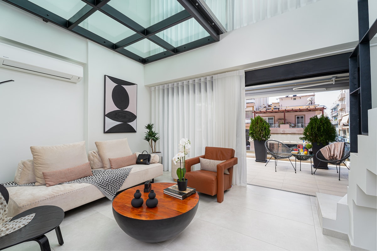 A modern living area is presented with a light-colored sofa and an armchair in brown. A circular coffee table holds decorative items. Large glass doors open to a balcony, allowing natural light to fill the space. White sheer curtains provide privacy while maintaining brightness.