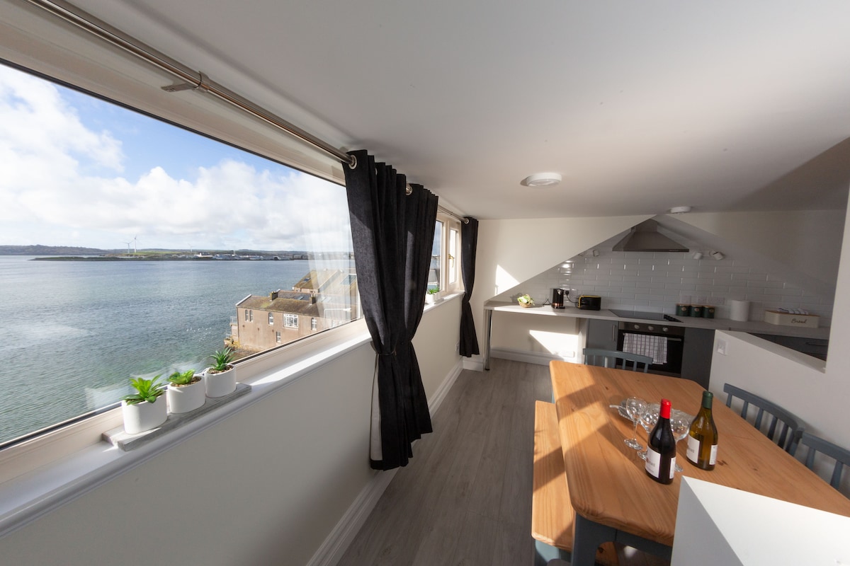 An airy dining area features a wooden table set for a gathering, complemented by two wine bottles and glasses. Large windows frame the serene water views, allowing natural light to illuminate the space. Minimalist decor emphasizes the coastal ambiance of this modern kitchen.
