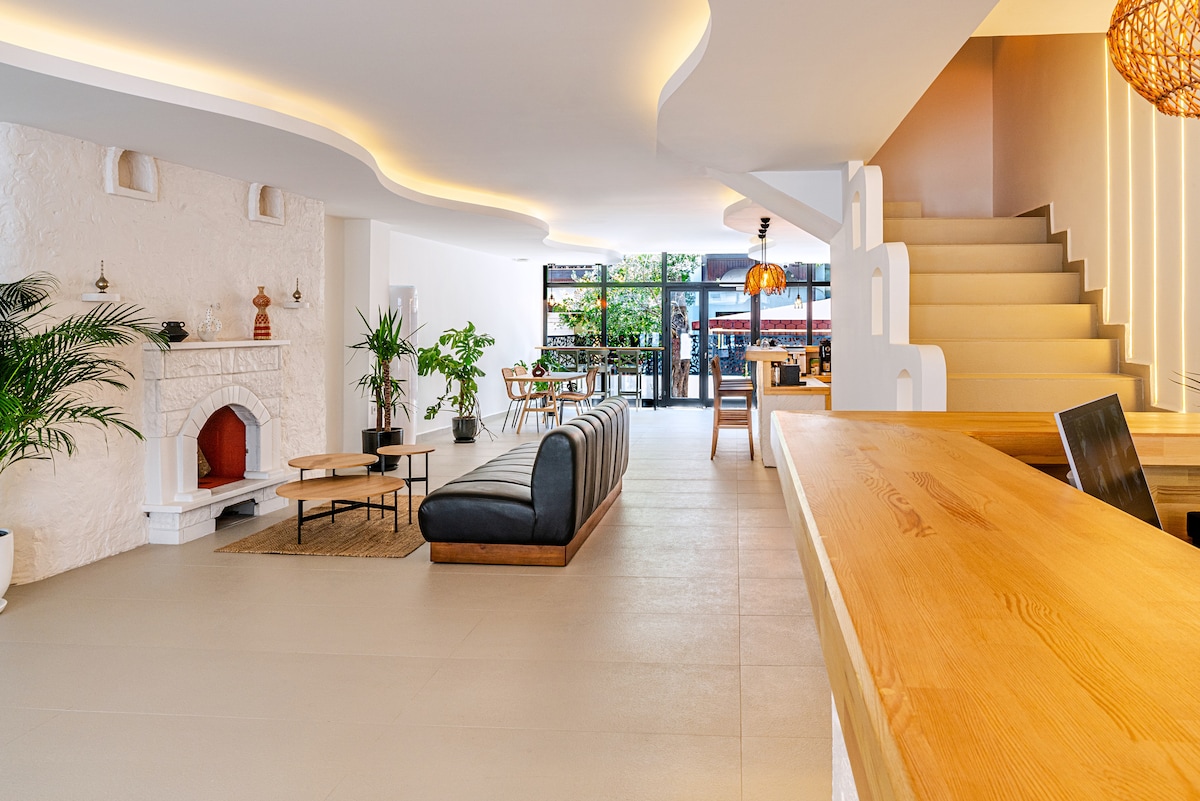 An open and airy lounge area features a modern seating arrangement with a long, curved sofa and wooden coffee tables. A white fireplace offers a focal point, while plants add a touch of greenery. Natural light streams through large windows, and a dining area is visible in the background.