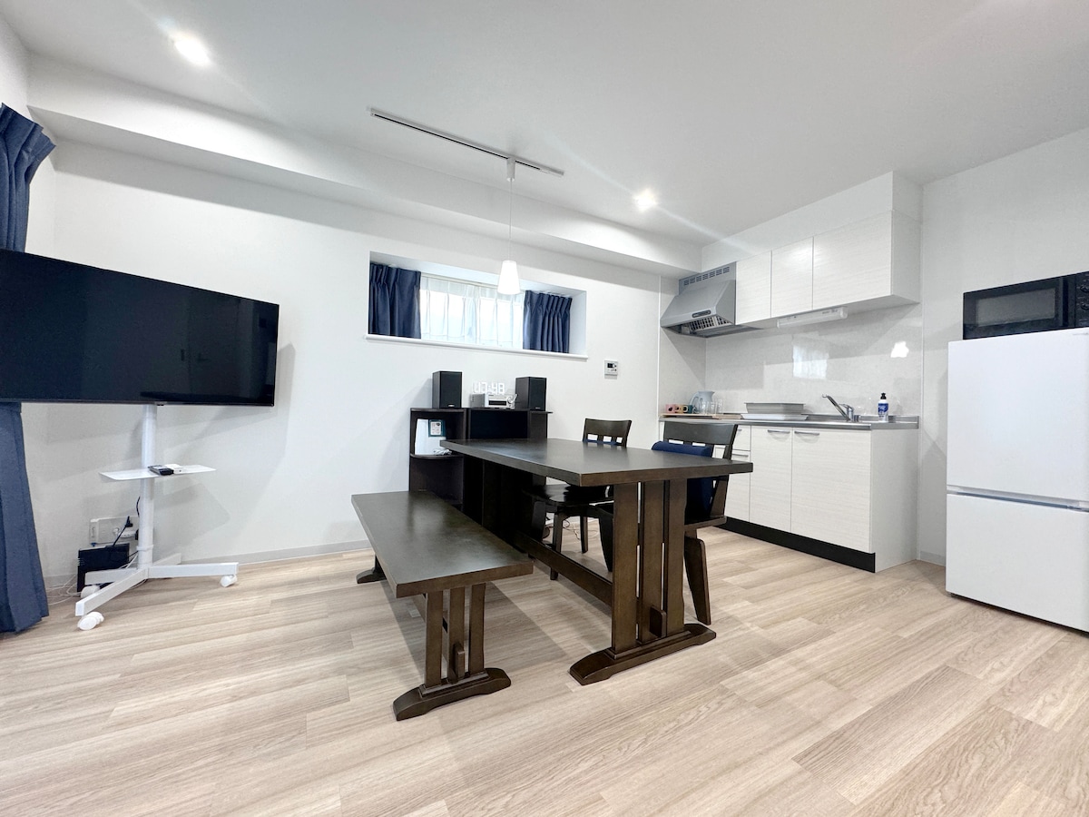 A modern kitchenette is paired with a dining area, featuring a large wooden table and sleek chairs. The space is brightened by natural light entering through a window, while a flat-screen TV is positioned for viewing. Clean, minimalistic design elements create a functional environment.