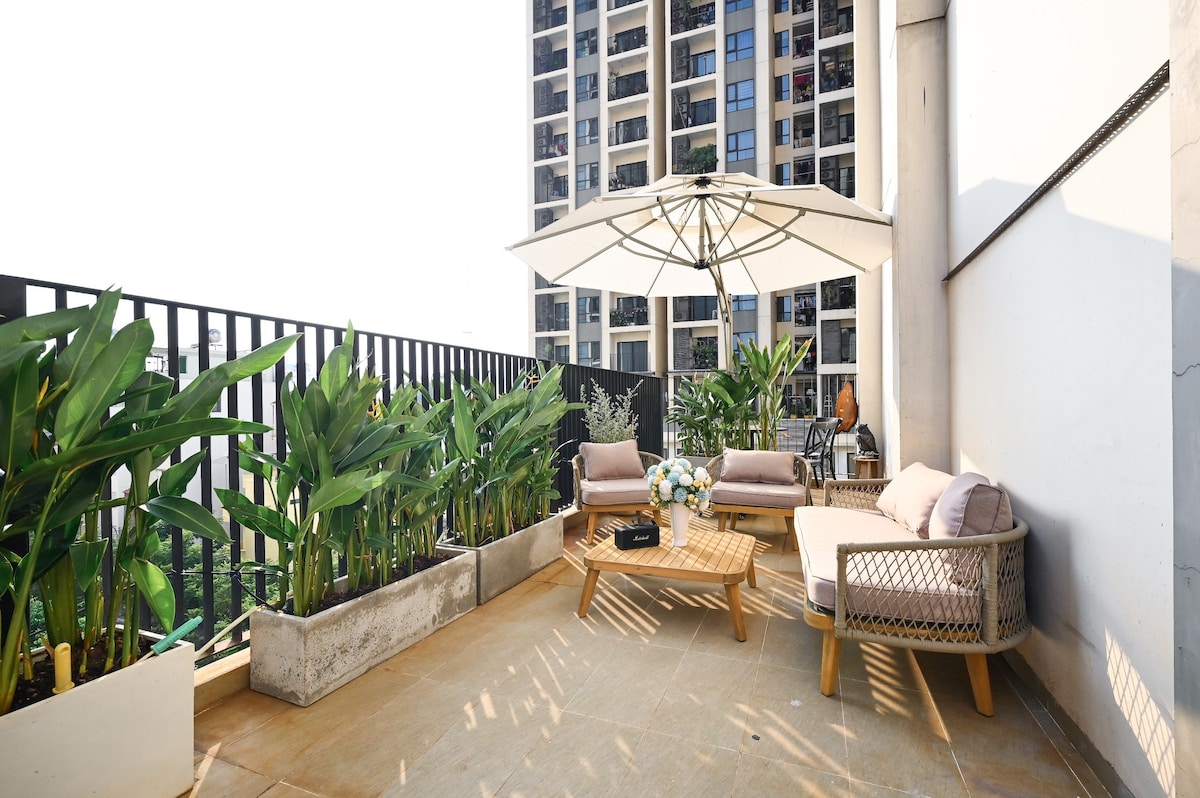 An outdoor terrace is shown, featuring a seating area with a round table and two sofas. Potted plants line the perimeter, adding greenery. A large umbrella provides shade, while a cityscape is visible in the background, indicating an urban environment.