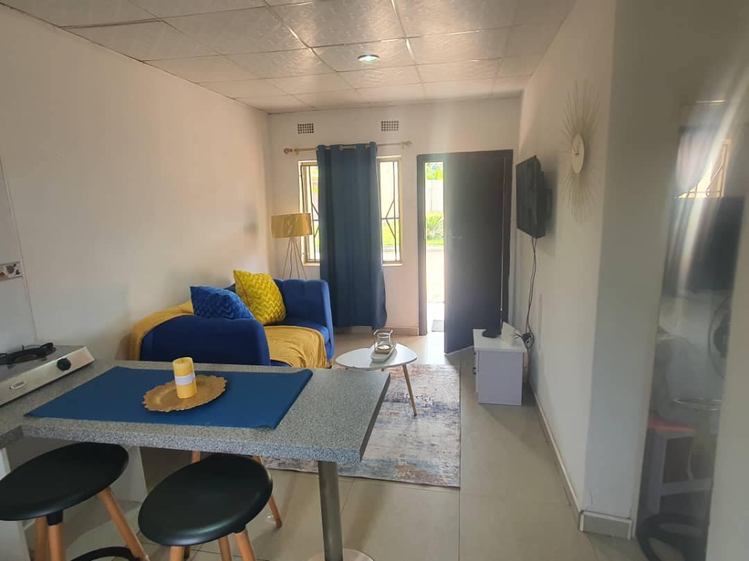 A compact living space features a blue and yellow sofa, accompanied by a small round table with bar stools. A flat-screen television is mounted on the wall, and sunlight enters through windows adorned with blue curtains. A neutral palette enhances the overall ambiance.