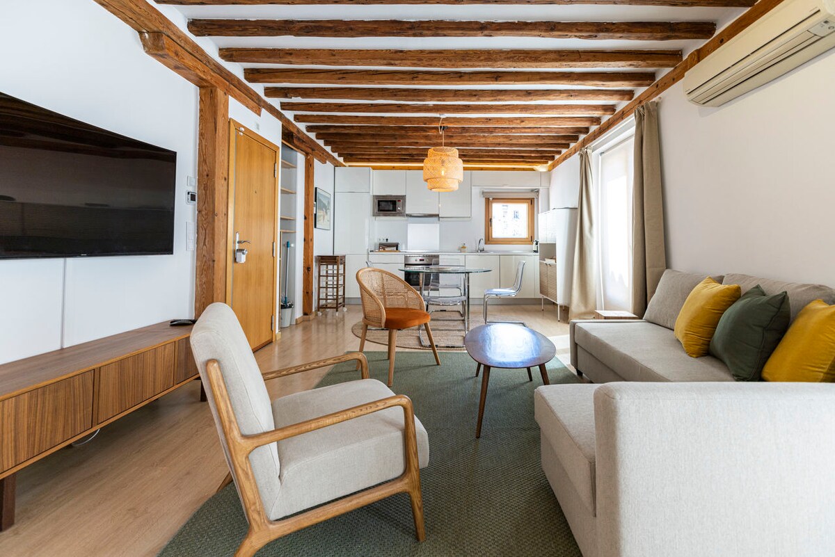 The living area features a light-colored sofa accompanied by a matching armchair and a round coffee table. A low wooden TV console holds a flat-screen television. Exposed wooden beams adorn the ceiling, and bright natural light enters through windows, enhancing the space.