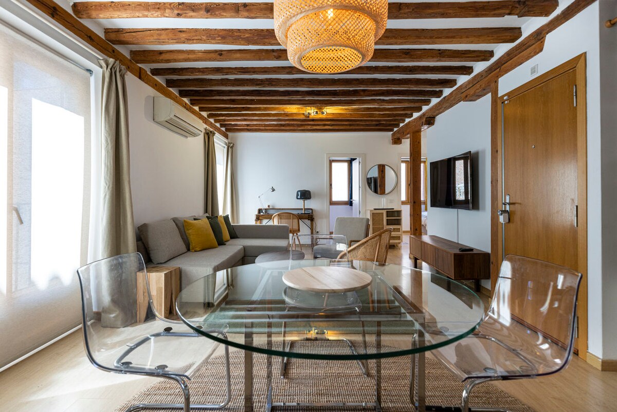 A spacious living area features a round glass dining table surrounded by transparent chairs. Natural light streams through large windows, highlighting the wooden beams on the ceiling. A comfortable sofa, wooden furniture, and a stylish chandelier enhance the inviting ambiance.