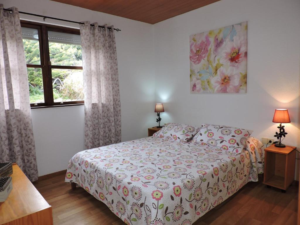 A comfortable sleeping area features a spacious bed adorned with a floral patterned duvet. Two bedside lamps provide soft lighting, and sheer curtains frame a window, allowing natural light to brighten the space. A piece of floral artwork is displayed above the bed.