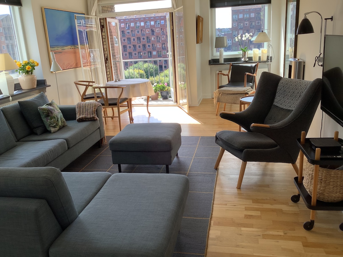 A light-filled living space features modern furnishings, including a gray sectional sofa, an accent chair, and a stylish ottoman. Large windows provide a view of the outdoor balcony, while a dining table and chairs are positioned in the background, inviting relaxation.