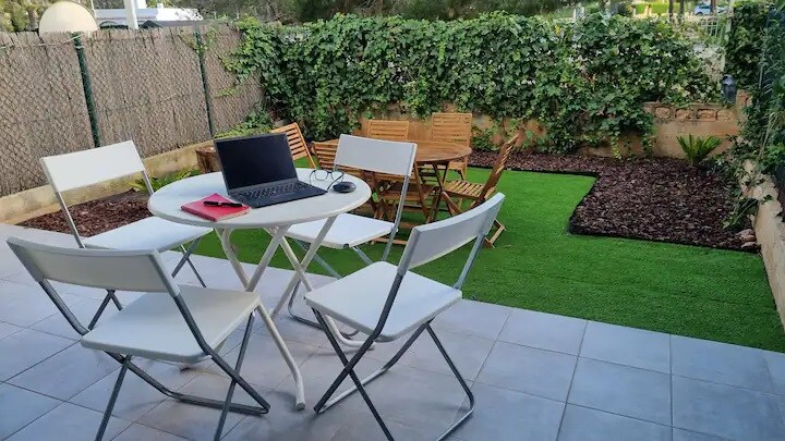 An outdoor seating area is visible, featuring a round table surrounded by four white chairs. A wooden dining table with matching chairs is seen in the background. Lush green plants and landscaping create a natural atmosphere, enhancing the space.
