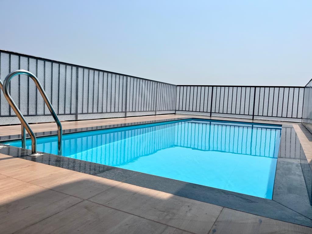 A rooftop swimming pool offers a refreshing retreat, surrounded by clear blue water and sleek stone flooring. Metal railings provide safety while showcasing an unobstructed view of the sky, creating a serene space for relaxation and enjoyment.