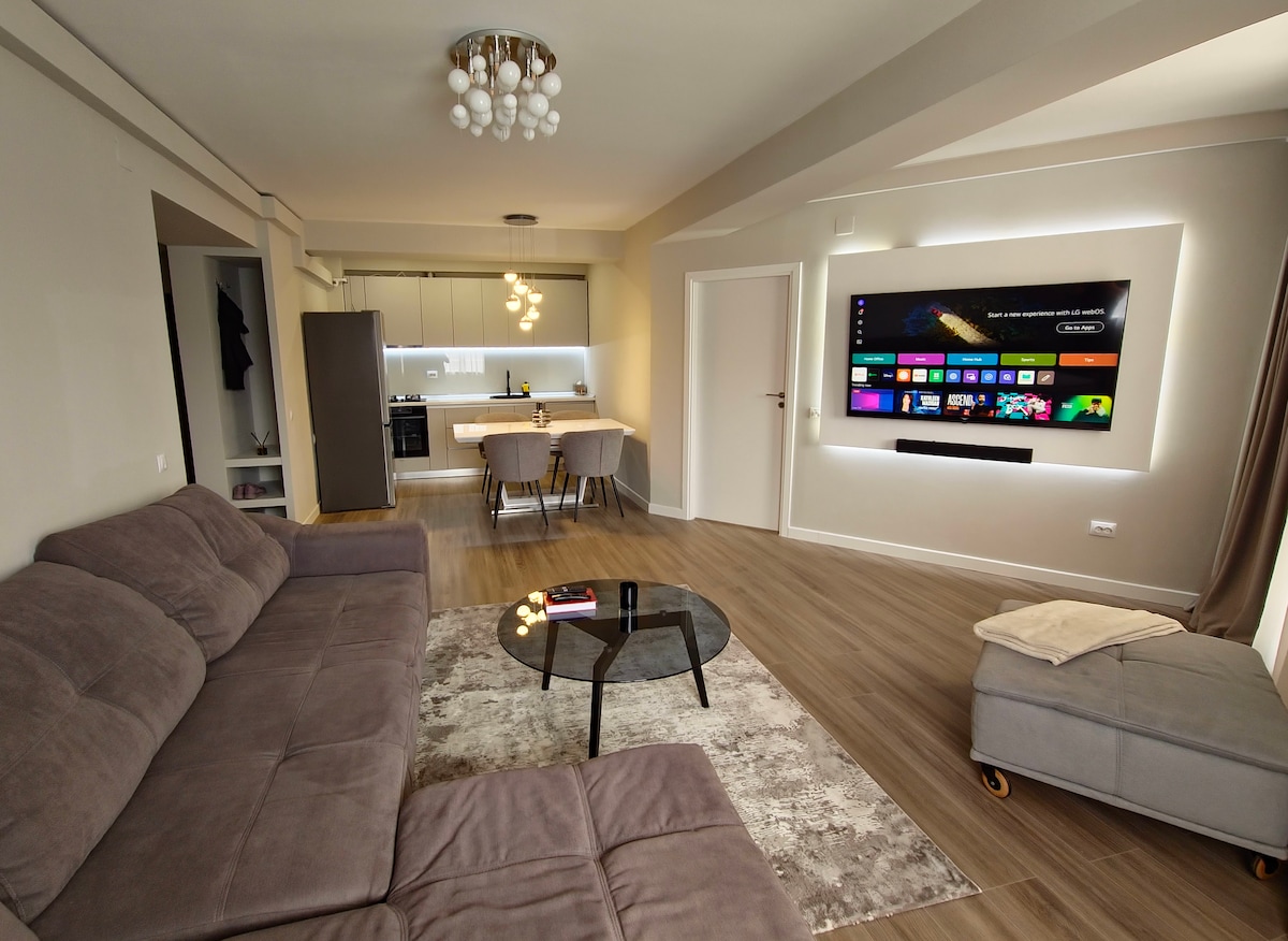 A modern living area is presented with a spacious, light grey sofa and a round glass coffee table atop a soft area rug. A wall-mounted TV is visible, surrounded by subtle ambient lighting. A dining space with modern chairs is situated near a sleek kitchen.