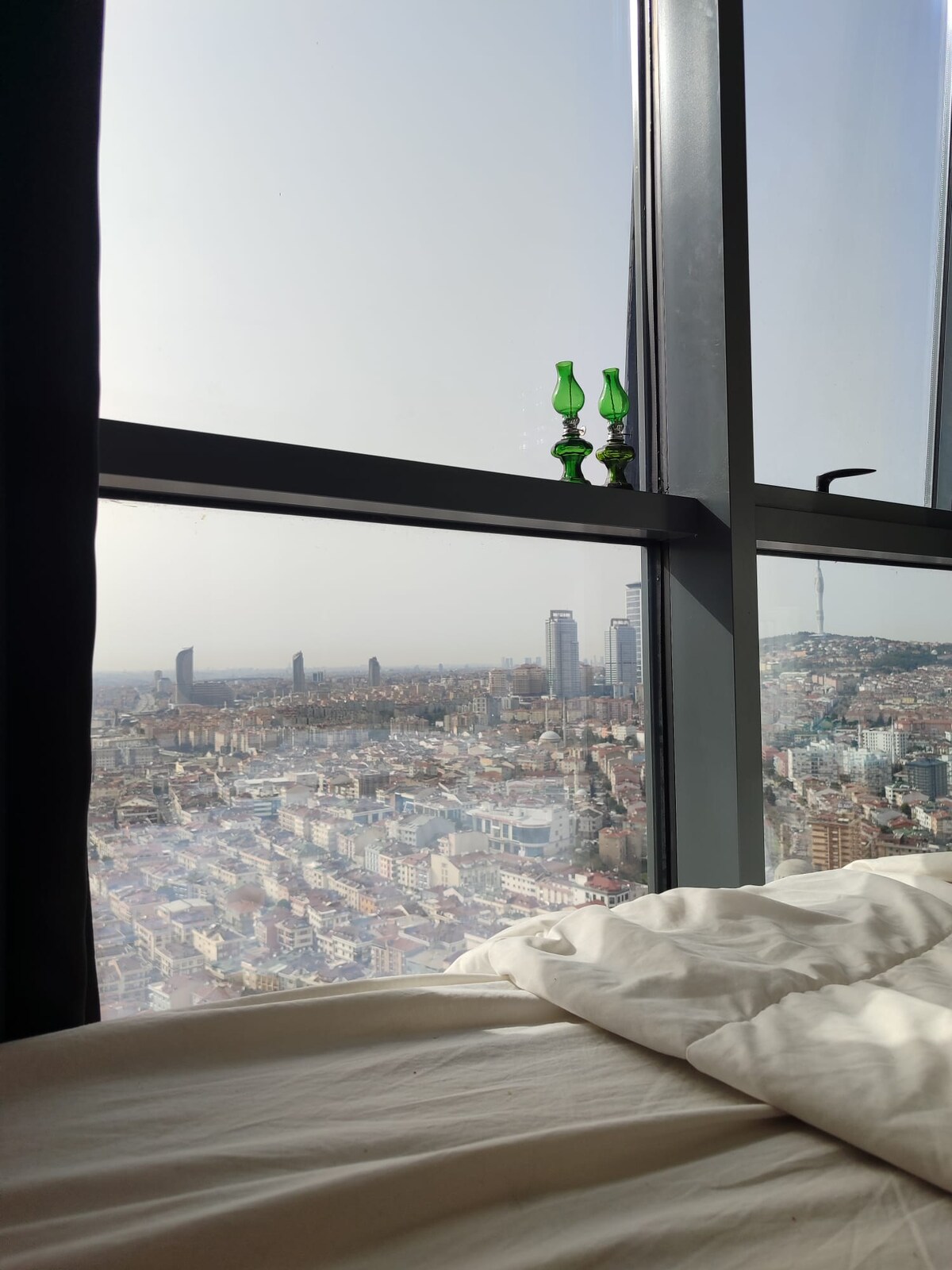 A stunning city view is captured through expansive glass windows. Two green glass decorative pieces rest on the windowsill, complementing the urban landscape below. Soft bedding is visible, creating a relaxed setting that invites guests to unwind while enjoying the scenic panorama.