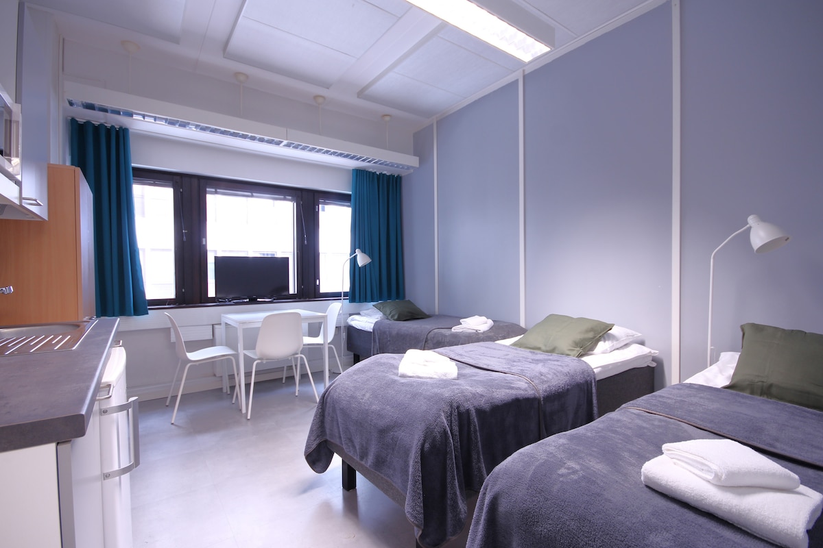 A well-organized room features three separated beds, each with plush blankets and neatly folded towels. A small, simple desk and chair set are positioned near the window, which is dressed with dark curtains. Natural light brightens the space, complemented by light gray walls.