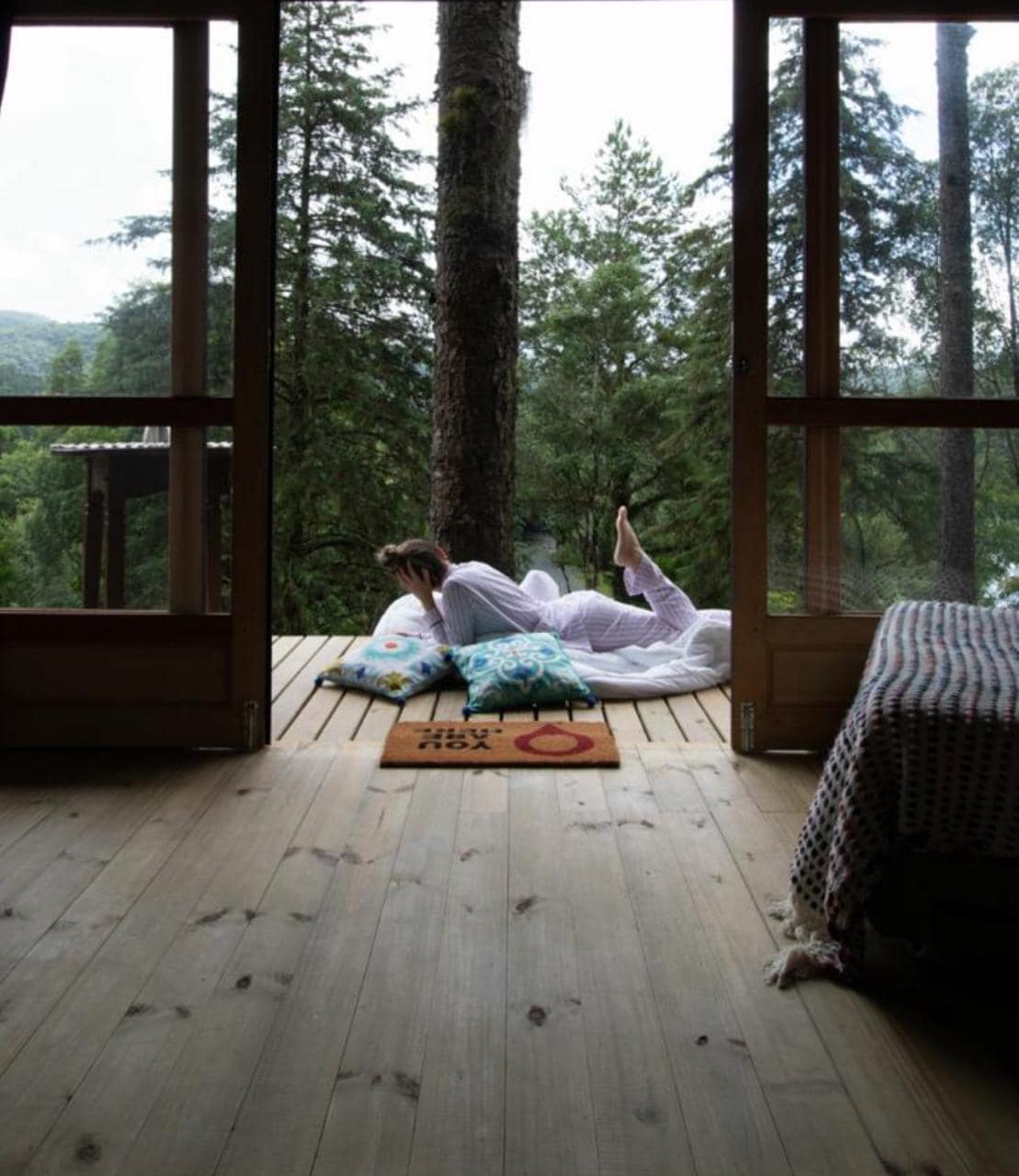 A serene cabin interior is observed, featuring a large open doorway that invites natural light. Cozy cushions are placed on a wooden deck, nestled amongst lush greenery. The floor showcases natural wood textures, enhancing the tranquil atmosphere of the space.