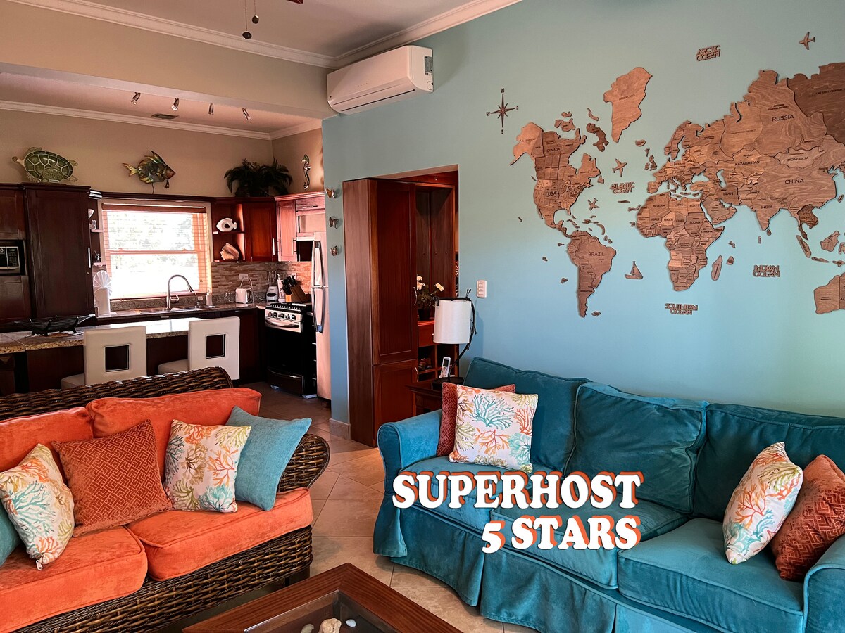 A welcoming living area features comfortable couches in vibrant orange and blue, accented with decorative pillows. A world map adorns the wall, adding a global touch. The background reveals a modern kitchen with wooden cabinetry and stainless steel appliances.