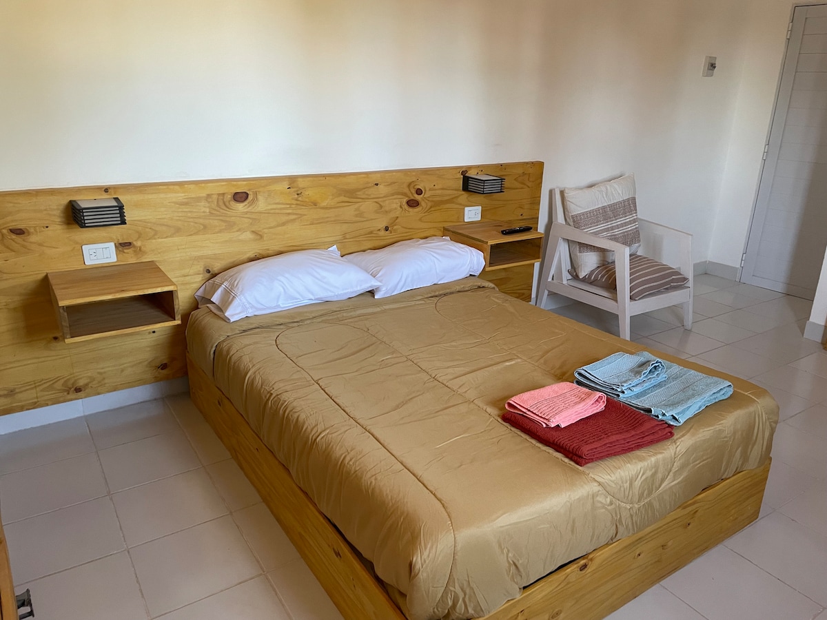 A simple bedroom features a spacious wooden bed with a neutral bedspread. Two neatly arranged towels in pink and blue are placed at the foot of the bed. Light-colored walls and tiled flooring contribute to a bright and clean atmosphere. A minimalist chair is positioned nearby.
